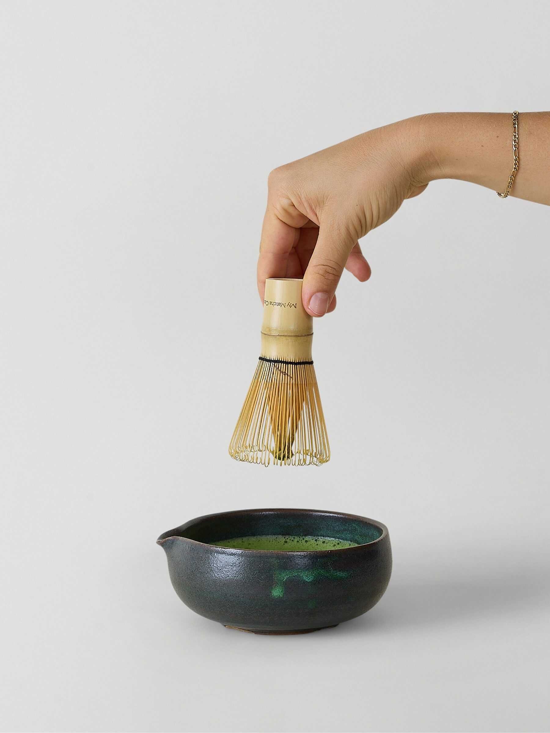 Person using a bamboo whisk to stir green tea in a black ceramic bowl on a light gray background