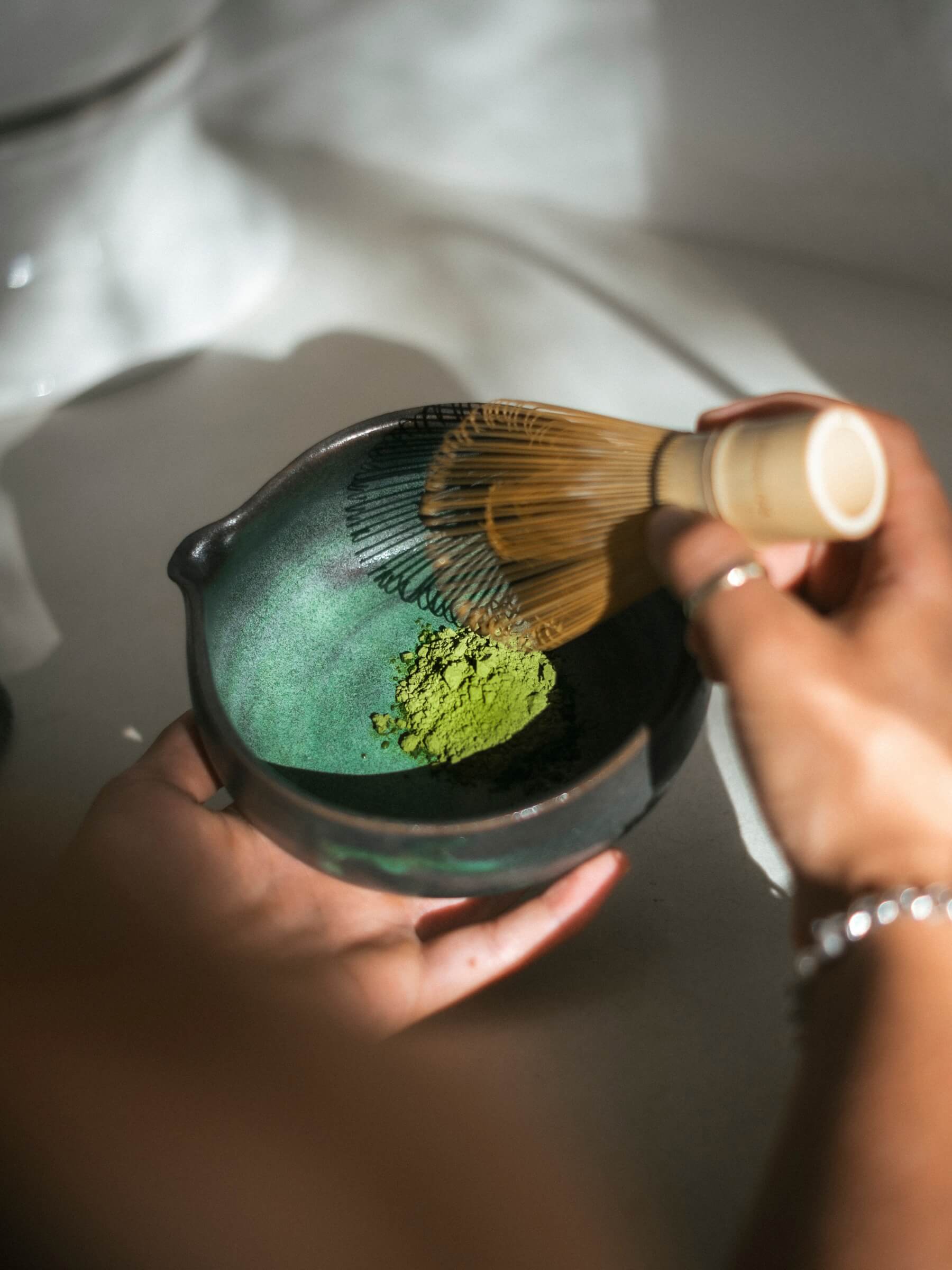 Vibrant green matcha powder in a ceramic blue being held in the sun with a bamboo whisk